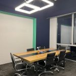 Interior view of a modern corporate meeting room containing a long rectangular wooden table surrounded by black wheeled office chairs, illuminated by two large square geometric pendant lights mounted on a dark grey ceiling, with a white wall panel and a window in the background