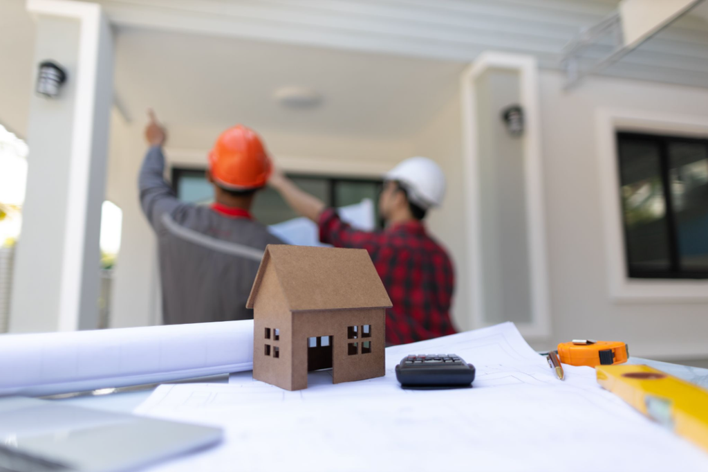 A small cardboard model house resting on architectural blueprints alongside a calculator and measuring tape, with construction workers inspecting a home in the background.