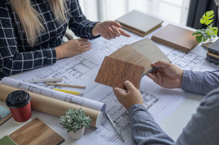 Engineers holding wood flooring material samples over architectural blueprints spread on a desk during a consultation meeting.