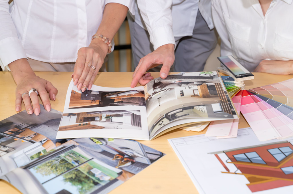 Interior designers reviewing home design magazine pages and color swatch fan decks spread across a wooden table next to architectural blueprints.