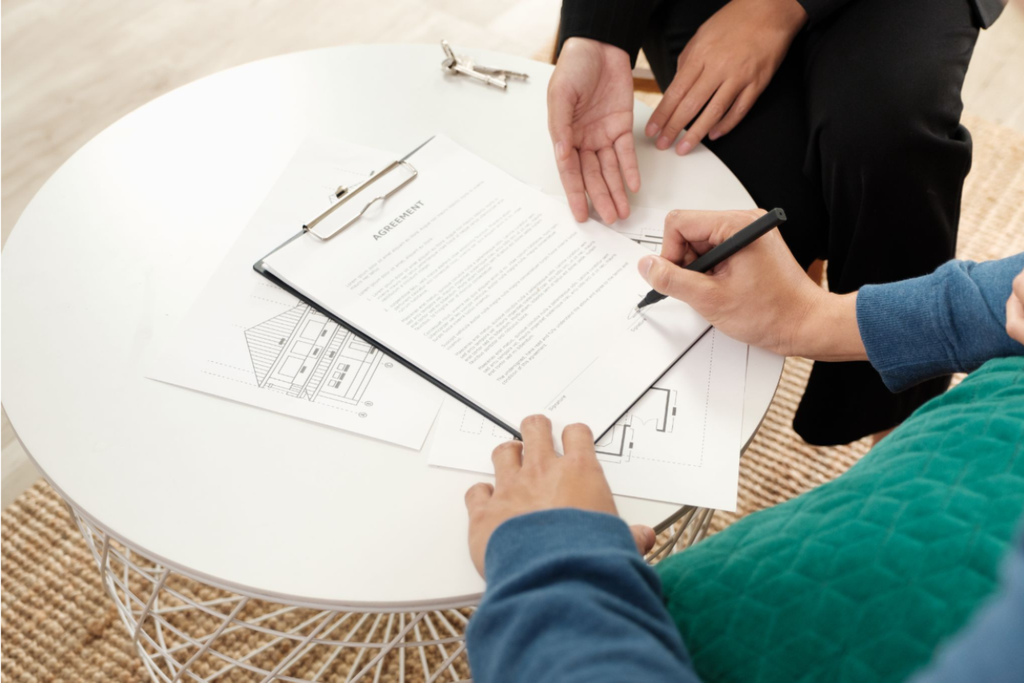 A person signing a real-estate agreement on a clipboard resting on a white coffee table alongside architectural blueprints.