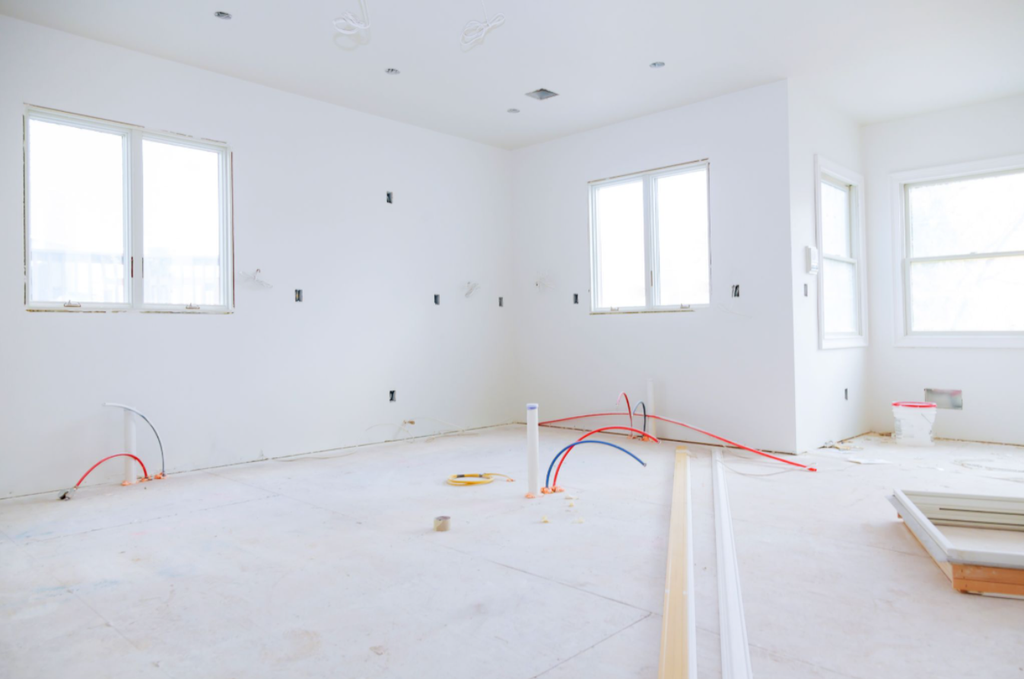 An unfinished room undergoing home renovation, showing white walls with exposed electrical outlets, protruding plumbing pipes on the floor, and windows.